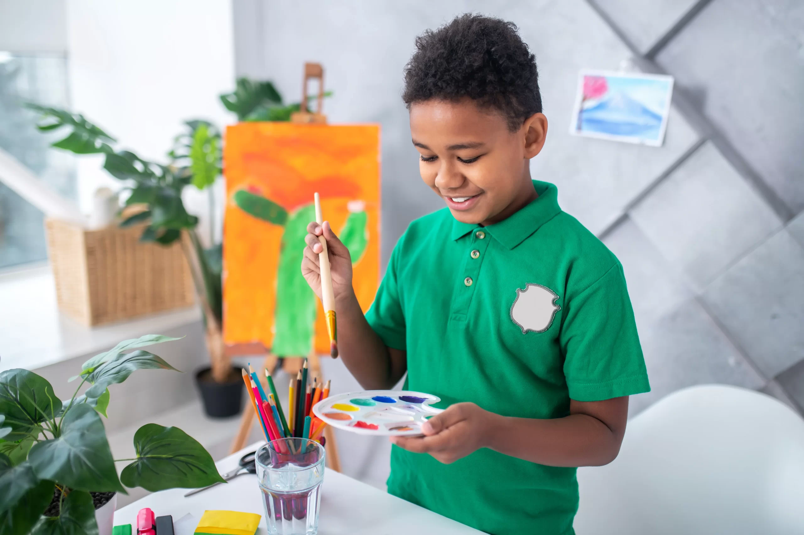 boy of primary school age in green tshirt holding paintbrush palette with paints